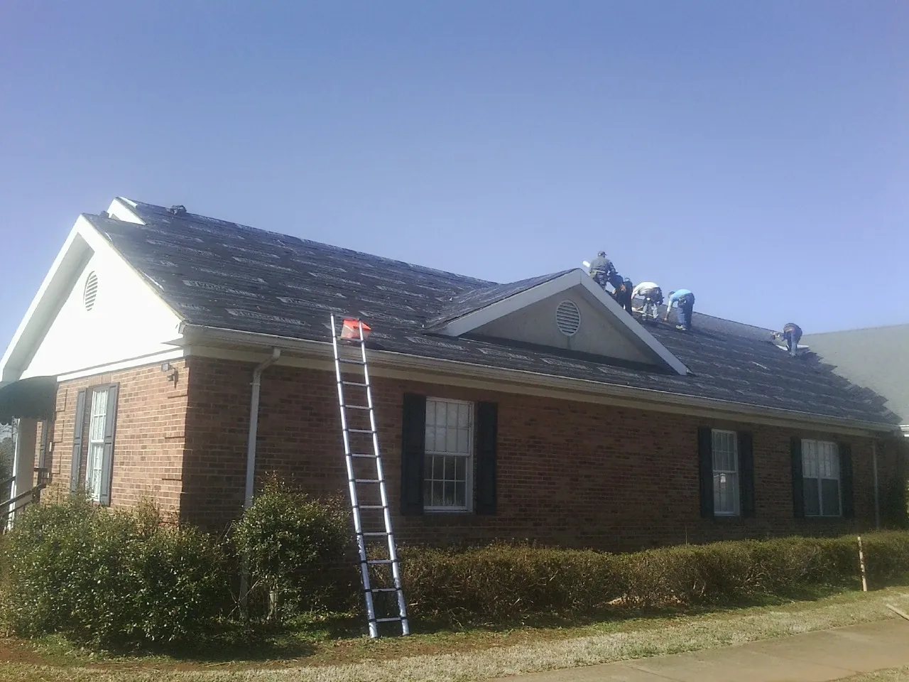 Crew tearing shingles off a brick ranch roof with a ladder set — Emergency Roof Repair work by our crew in Hospers