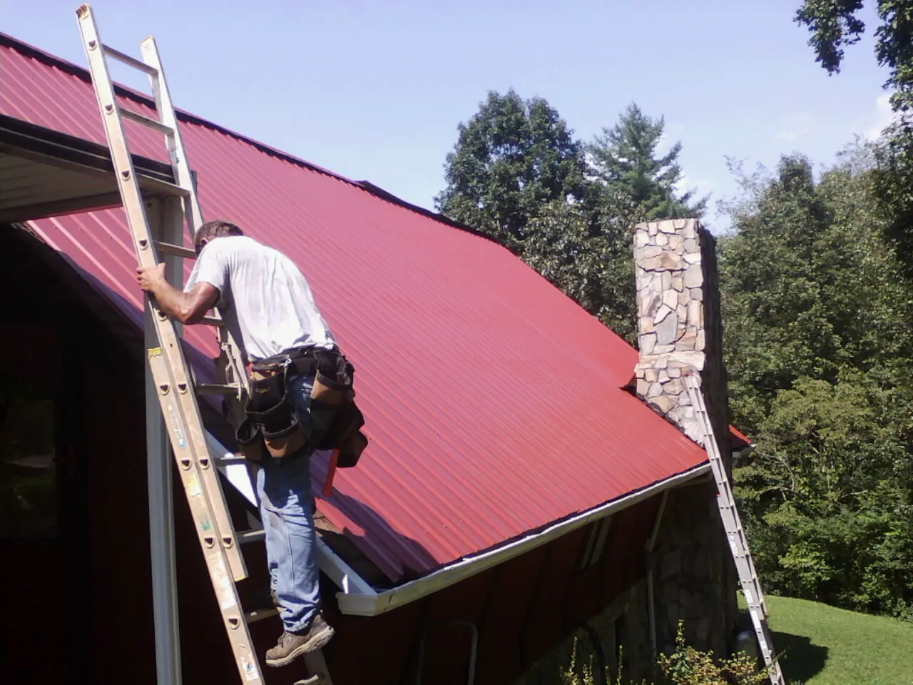 Roofer on a ladder beside a red metal roof and stone chimney — Soffit & Fascia Repair by Roofing Brothers in Hospers